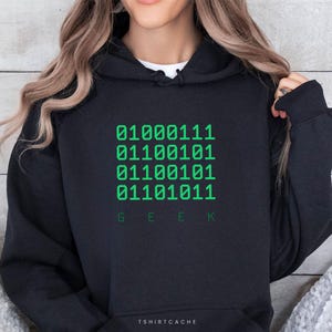 May include: Black hoodie with a green binary code design and the word "GEEK" in green at the bottom. The hoodie has a drawstring hood and a front pocket. The text "TSHIRTCACHE" is at the bottom.