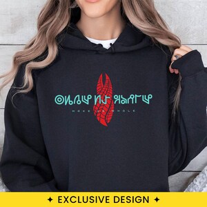 May include: A black hooded sweatshirt featuring a red graphic of a stylized, segmented vertical shape centered on the chest. Above the graphic is light blue alien-style text, and below it is the white text "MAKE US WHOLE" in a sans-serif font.