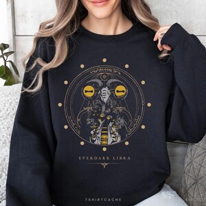 May include: Black sweatshirt with a gold and gray graphic design. The design features a circular illustration with a horned figure, symbols, and the text "EVERDARK LIBRA". The brand name "TSHIRTCACHE" is at the bottom.