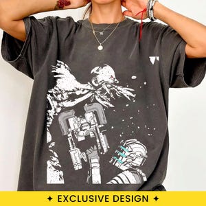 May include: A charcoal gray short-sleeve t-shirt featuring a white graphic print of a sci-fi character wearing a helmet with blue light accents, facing a large, jagged, monstrous creature with red eyes and sharp, protruding teeth and claws.