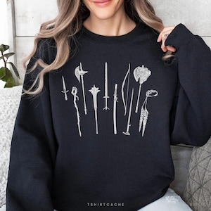 May include: A black long-sleeve sweatshirt featuring a white line art graphic of various medieval weapons, including a dagger, mace, sword, katana, bow, and axe. The text TSHIRTCACHE is printed in small white letters at the bottom center.