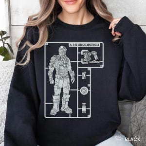 May include: A black long-sleeve sweatshirt featuring a light gray graphic print of a model kit layout. The design includes a character in armored gear labeled A 1:10 ISSAC CLARKE RIG L3, alongside various equipment components numbered one through five.