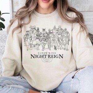 May include: Cream-colored sweatshirt featuring a black line drawing of multiple characters from the video game Elden Ring. The text "ELDEN RING NIGHT REIGN" is printed below the illustration. The sweatshirt has a crew neck and long sleeves.