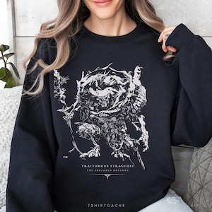May include: A black crewneck sweatshirt featuring a white graphic illustration of a skeletal, horned creature holding a staff. Below the creature, the text reads: TRAITOROUS STRAGHESS THE FORSAKEN HOLLOWS. The bottom of the shirt displays TSHIRTCACHE.