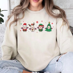 May include: Cream-colored sweatshirt featuring four cartoon characters dressed in Christmas attire. The characters include a Santa, a reindeer, an elf, and a Christmas tree, each with bat wings. The text "TSHIRTCACHE" is at the bottom.