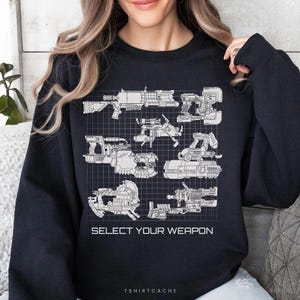 May include: A black long-sleeve sweatshirt featuring a white grid pattern graphic with various sci-fi weapon illustrations. Below the weapons, the text reads "SELECT YOUR WEAPON" in a clean, sans-serif font. The design is centered on the chest.