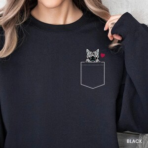 May include: A black crewneck sweatshirt featuring a white outline of a chest pocket. Peeking out from the pocket is a grey, furry creature with a black visor and a small red heart graphic positioned to the right of the creature's head.