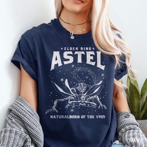 May include: Navy blue t-shirt featuring a white graphic of a skeletal creature with wings and the text "ELDEN RING ASTEL NATURALBORN OF THE VOID". The design includes stars and a celestial theme.