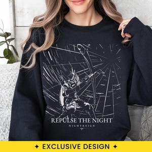 May include: A black long-sleeve sweatshirt featuring a white graphic print of a figure holding a bow and arrow amidst radiating lines. The text "REPULSE THE NIGHT" and "NIGHTREIGN" is printed in white below the central illustration on the chest.