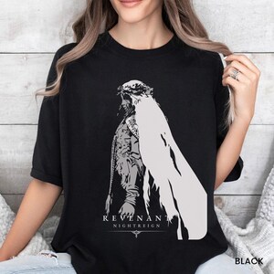 May include: A black short-sleeved t-shirt featuring a large, light gray graphic print of a figure in profile wearing a veil and crown. Below the figure, the text reads REVENANT NIGHTREIGN in a clean, sans-serif font with a small decorative icon.