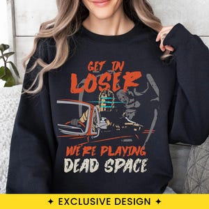 May include: A black long-sleeve sweatshirt featuring a graphic print of a character in a helmet driving a car with a creature in the passenger seat. The text reads GET IN LOSER WE'RE PLAYING DEAD SPACE in orange and white distressed lettering.
