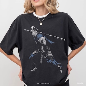 May include: A black short-sleeved t-shirt featuring a graphic print of a knight in armor holding a long sword. The illustration uses white, grey, and blue tones to depict the figure in a dynamic stance. The shirt has a crew neckline.