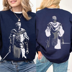 May include: A navy blue crewneck sweatshirt featuring a graphic illustration of a character in a hooded cloak and layered tunic. The front shows the character facing forward holding a staff, while the back shows the character from behind.