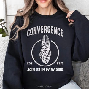 May include: A black long-sleeve sweatshirt featuring a white distressed graphic print. The design includes the text CONVERGENCE, a central stylized marker symbol inside a circle, the text EST 2215, and the phrase JOIN US IN PARADISE.