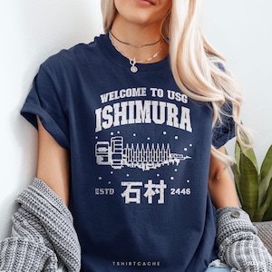 May include: A navy blue short-sleeve t-shirt featuring a white distressed graphic print that reads WELCOME TO USG ISHIMURA, with a stylized spaceship illustration, the Japanese characters for Ishimura, and the text ESTD 2446 centered on the chest.