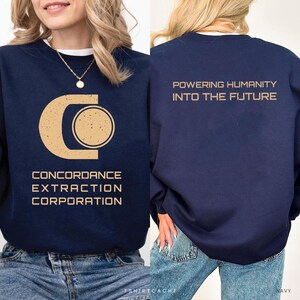May include: A navy blue crewneck sweatshirt featuring a distressed tan graphic on the front and back. The front displays a stylized C logo with the text CONCORDANCE EXTRACTION CORPORATION. The back features the text POWERING HUMANITY INTO THE FUTURE.