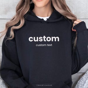 以下が含まれることがあります： 白い文字で「custom」と書かれた黒いパーカー。「custom text」というテキストが下にあります。パーカーには、ドローストリング付きのフードとフロントポケットが付いています。