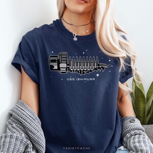 May include: Navy blue t-shirt featuring a pixelated graphic of a spaceship with the text "USG ISHIMURA" in white. The design includes stars and a futuristic aesthetic. The shirt has rolled-up sleeves.