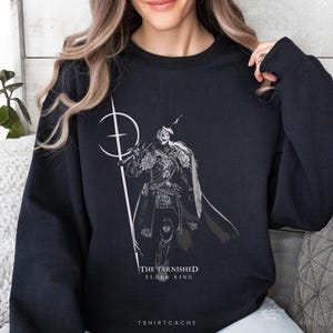 May include: Black sweatshirt featuring a detailed illustration of a warrior holding a spear, with the text "THE TARNISHED ELDEN RING." The design is in shades of gray and white. The brand name "TSHIRTCACHE" is printed at the bottom.