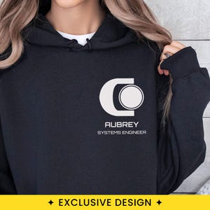 May include: A black hooded sweatshirt featuring a white graphic on the chest. The design includes a stylized letter C logo with a circle inside, followed by the text AUBREY and the words SYSTEMS ENGINEER printed in a clean, sans-serif font.