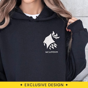 May include: A black hooded sweatshirt featuring a white graphic on the left chest. The design depicts a stylized, textured silhouette of a head with flowing hair or flames, positioned above the text MCLOVIN99 in a clean, sans-serif font.