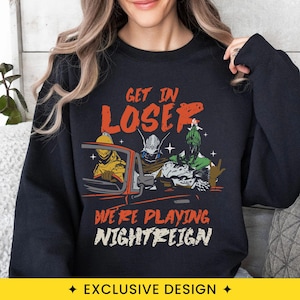 May include: A black long-sleeve sweatshirt featuring a graphic print of three armored figures in a red convertible car. The text reads GET IN LOSER WE'RE PLAYING NIGHTREIGN in orange and white lettering. The design includes small white star accents.