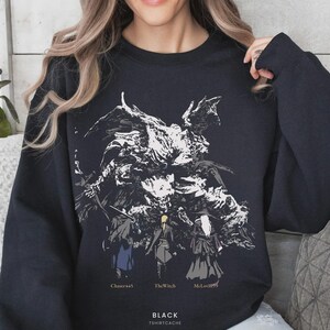 May include: A black long-sleeve sweatshirt featuring a large white graphic of a dark, winged creature looming over three small figures. Below the figures, the text reads Chaser445, TheWitch, and McLovin99, with BLACK TSHIRTCACHE at the bottom.
