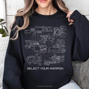 May include: A black long-sleeve sweatshirt featuring a white grid pattern graphic with various technical weapon illustrations and the text "SELECT YOUR WEAPON" printed in white capital letters across the center of the chest area.