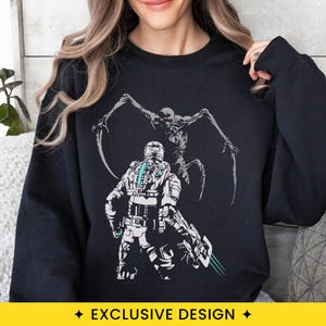 May include: A black long-sleeve sweatshirt featuring a graphic print of a futuristic armored character in a helmet with a glowing blue spine light, facing a tall, spindly, multi-limbed creature with sharp claws and glowing red eyes.