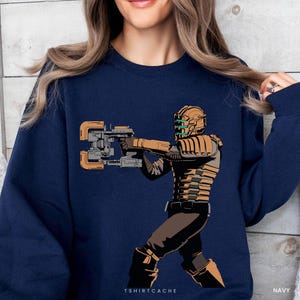 May include: A navy blue crewneck sweatshirt featuring a graphic illustration of a character in a tan and brown armored suit holding a large mechanical tool. The character wears a helmet with glowing blue lights. The text TSHIRTCACHE is printed below.