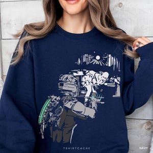 May include: Navy blue sweatshirt featuring a graphic design of a futuristic figure with a weapon and a mechanical arm. The design is in shades of gray, white, and green. The text "TSHIRTCACHE" is printed at the bottom.