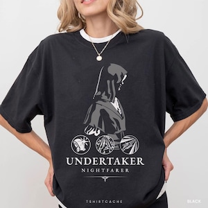 May include: Black t-shirt featuring a graphic of a cloaked figure with the text "UNDERTAKER NIGHTFARER". Below the text are three circular designs. The shirt also has the text "TSHIRTCACHE" and "BLACK".