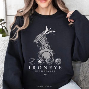 May include: A black long-sleeve sweatshirt featuring a light gray graphic of a hooded figure in profile. Below the figure are four circular icons and the white text "IRONEYE NIGHTFARER" with a small decorative line element underneath the text.