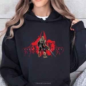 May include: Black hoodie featuring a red graphic of a futuristic soldier with a weapon, set against a red abstract background. The text "FUCK YOU MARKER" is printed below the graphic. The brand name "TSHIRTCACHE" is printed at the bottom.