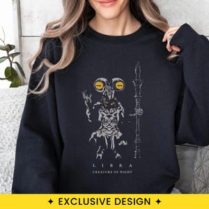 May include: A black crewneck sweatshirt featuring a graphic print of a horned creature with large yellow eyes holding a staff. The design includes the text LIBRA CREATURE OF NIGHT in white lettering below the illustration of the creature.