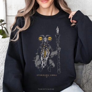 May include: Black sweatshirt featuring a detailed illustration of a creature holding a staff. The design includes yellow and gray accents, with the text "EVERDARK LIBRA" and "TSHIRTCACHE" printed below the graphic.