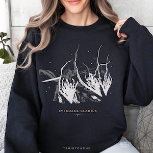 May include: Black sweatshirt featuring a white and gray graphic design of a stylized creature with a sword. The text "EVERDARK GLADIUS" is printed below the graphic, with the text "TSHIRTCACHE" at the bottom.