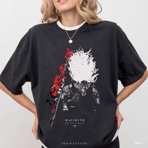 May include: A black short-sleeve t-shirt featuring a graphic print of a creature with a large white textured mane and red accents. Below the graphic, the text reads MALIKETH THE BLACK BLADE. The shirt has a crew neckline and a relaxed fit.