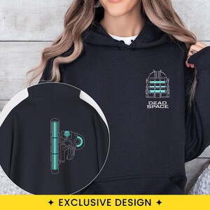 May include: A black hoodie featuring a white and teal Dead Space logo on the chest. An inset circle shows the back of the hoodie, which displays a vertical teal light bar graphic with mechanical details and a circular element on the right side.
