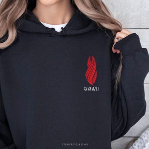 May include: Black hoodie with a red graphic design on the chest. The design is a stylized, flame-like shape with intricate detailing. The text "TSHIRTCACHE" is printed at the bottom.