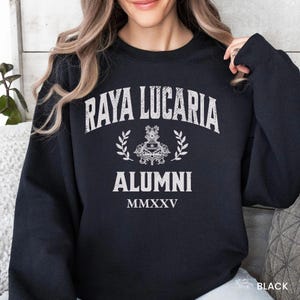 May include: A black long-sleeve sweatshirt featuring white distressed-style text that reads Raya Lucaria Alumni with a central crest graphic flanked by laurel branches and the Roman numeral MMXXV printed below the text in a clean font.