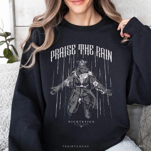 May include: Black sweatshirt featuring the phrase "PRAISE THE RAIN" in white gothic lettering above a graphic of a character with outstretched arms. The graphic includes the text "NIGHTREIGN ELDEN RING" and "TSHIRTCACHE".