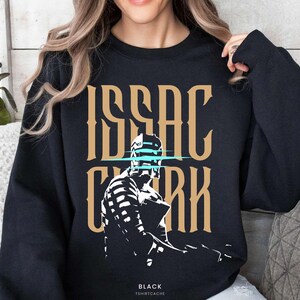May include: Black sweatshirt featuring the name "ISAAC CLARK" in a gold, stylized font, with a blue horizontal line through the middle. Below the text is a white and black graphic of a character. The word "BLACK" and "TSHIRTCACHE" are printed at the bottom.