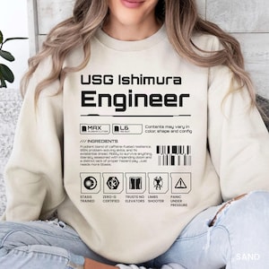 May include: A sand-colored crewneck sweatshirt featuring black graphic text that reads USG Ishimura Engineer. The design includes a barcode, technical icons, and humorous ingredient text referencing caffeine, problem-solving, and existential dread.