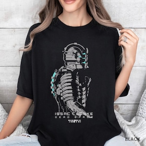 May include: A black t-shirt featuring a graphic print of the character Isaac Clarke from Dead Space in profile. The design uses grey and white lines with cyan accents on the helmet and suit. The text reads ISSAC CLARKE DEAD SPACE.