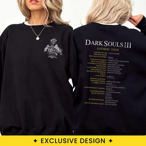 May include: A black long-sleeve sweatshirt featuring a grey graphic of a knight on the front chest. The back displays the white text DARK SOULS III LOTHRIC TOUR followed by a list of locations and boss names in white and gold lettering.