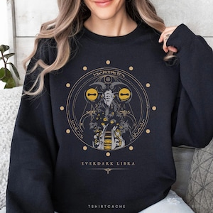 May include: Black sweatshirt with a gold and white graphic design. The design features a horned figure within a circular frame, with the text "EVERDARK LIBRA" below. The brand name "TSHIRTCACHE" is at the bottom.