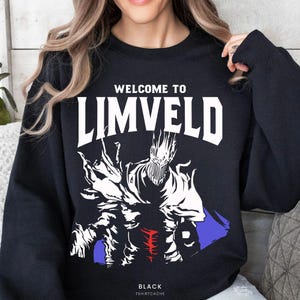 May include: Black sweatshirt with the text "WELCOME TO LIMVELD" in white block letters. Below the text is a white and blue graphic of a creature. The word "BLACK" and "TSHIRTCACHE" are printed at the bottom.