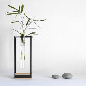 Op de afbeelding: Een zwarte metalen vaas met een doorzichtige glazen buis om een ​​enkele stengel groen te houden. De vaas staat op een wit oppervlak met twee grijze stenen.