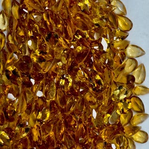 May include: A collection of faceted, teardrop-shaped gemstones in shades of yellow and amber. The gems are scattered across a white surface, creating a sparkling display. The gemstones are likely citrine, a popular choice for jewelry.